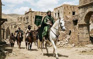A photograph of a group of seven horsemen in traditional, non-western clothing riding through the narrow, dusty street of an ancient stone village. The central figure rides a distinct white horse and wears a bright green robe with a green turban. The rider immediately behind him, on a brown horse, holds a dark banner with white Arabic text at the top and the English words "MU'MIN BELIEVERS" below it. The other riders are dressed in shades of brown, white, and black, and two riders to the left hold spears and shields. The surrounding buildings are constructed from rough, aged stone and sun-baked mud brick, with architectural details like a circular archway in the distance. The overall scene is set in a dry, barren, and sun-lit landscape under a blue sky with scattered clouds.