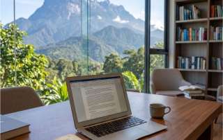 A photograph of a study desk overlooking Mount Kinabalu in Borneo. On the wooden table is an ancient, open manuscript with a magnifying glass resting on the text, beside a modern open laptop and copies of the books "DOGMA," using the provided cover designs.