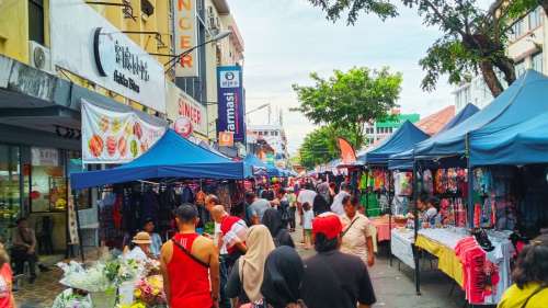 A bustling outdoor street market filled with shoppers, colorful vendor canopies, local crafts, and historic shoplots along Gaya Street in Kota Kinabalu.