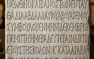 Close-up photograph of the authentic Hammat Gader inscription block, a dark volcanic basalt slab carved entirely with neat rows of ancient Greek block letters, beginning with a Christian cross in the top left corner.