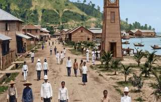 A vivid color reconstruction of early Jesselton (modern-day Kota Kinabalu) circa 1905. The image shows the iconic wooden Atkinson Clock Tower standing prominently near the sea, with the barren hills of Signal Hill in the background. Colonial officers in white uniforms and local residents in traditional attire walk along the unpaved waterfront road, capturing the resilient spirit of North Borneo’s early administrative capital.