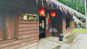 A child standing outside a traditional wooden Hakka House with a thatched roof and hanging red lanterns at the Sabah Museum Heritage Village in Kota Kinabalu.
