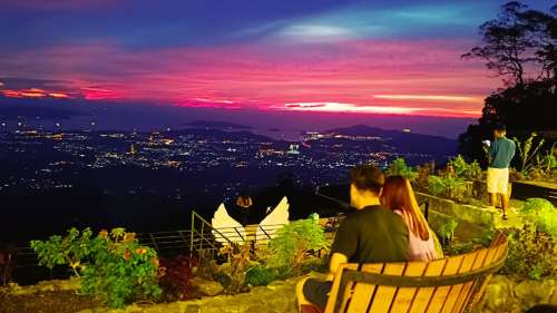 A couple sitting on a wooden bench at Kokol Hill overlooking a vibrant purple and crimson sunset sky over Kota Kinabalu city and the sea.