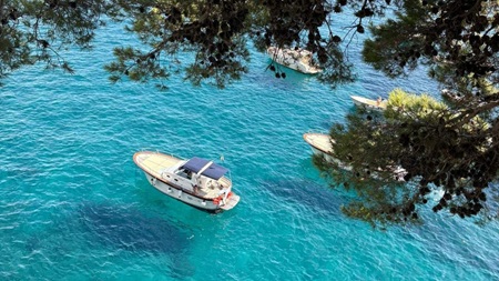 Modern day boats floating on clear turquoise water along the Amalfi Coast seen through green pine trees.