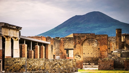 A landscape travel photograph capturing the modern peak of Mount Vesuvius standing behind the excavated stone columns and brick arches of the Pompeii ruins, ideal for readers of historical fiction author Don Peter.