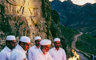 This portrait-oriented, mobile-optimized photograph captures a group of six Zoroastrian Mobeds (priests) performing a 'Yasna' ceremony or fire offering. They are dressed in immaculate white robes, symbolizing purity, and are clustered around a stone fire altar (an Atash Kadeh) built upon a cliffside overlooking a rugged mountain range in Iran. The central priest, a distinguished man with a grey beard, is actively feeding sandalwood and offerings into the intense flames using a metal ladle. The background is defined by massive, textured rock formations. Looming prominently on the cliff face is an ancient, large-scale relief carving of the Faravahar, the icon of Zoroastrianism representing Ahura Mazda and the guardian spirit. The sky above is a dramatic, clouded sunset, where powerful streaks of fire-orange and gold battle with deep purples and blues, visually representing the primal Zoroastrian theme of the cosmic struggle between truth (Asha) and falsehood (Druj). The composition is designed for mobile readability, using the vertical space to contrast the intimate human ritual with the vast historical and environmental elements.