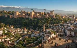 Digital historical artwork presenting a scenic view from an elevated plaza in the Albaicín quarter of Granada, Spain. Captures traditional whitewashed Moorish architecture, cobblestone streets, and cypress trees looking toward the Alhambra complex and snow-capped peaks of the Sierra Nevada.