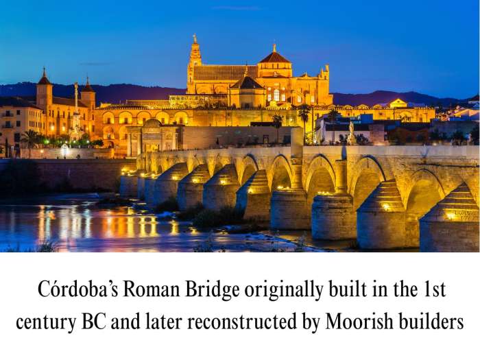 Vibrant twilight photograph showing the illuminated stone arches of the Roman Bridge of Córdoba glowing over the reflecting waters of the Guadalquivir River. In the background, the golden-lit Mosque-Cathedral (Mezquita), Puerta del Puente, and ancient city ramparts stand out dramatically against a deep blue evening sky.
