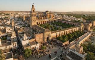 High-detail digital artwork showing an elevated aerial perspective of medieval Córdoba, Spain. Features the grand courtyard, bell tower, and cathedral structure of the Mezquita, flanked by the historic quarter, bustling plazas, and the ancient Roman Bridge over the river.