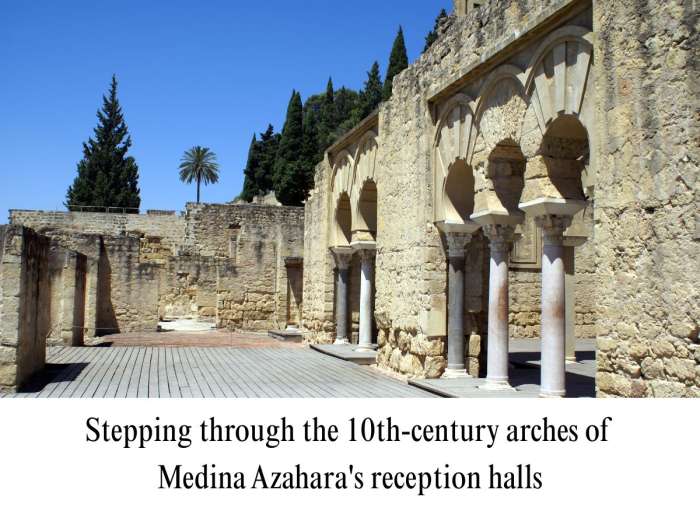 Photograph capturing a restored section of the Medina Azahara archaeological site near Córdoba, Spain. Focuses on a row of Caliphate horseshoe arches supported by white marble and gray stone columns, leading to a large open courtyard with timber decking. In the background are ancient retaining walls, tall cypress trees, a single palm tree, and a clear blue sky.