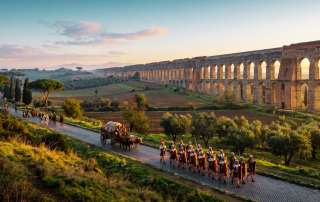 A sweeping, cinematic landscape reconstruction depicting the towering stone arches of the great Roman aqueducts alongside the Appian Way during the golden hour. This featured image anchors the historical guide, highlighting the scale of ancient imperial engineering, traditional Roman soldiers on the march, and the open countryside outside the city center.