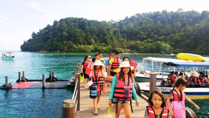 A lively group of tourists arriving at the Sapi Island wooden jetty pier in Tunku Abdul Rahman Marine Park. Wearing safety life jackets and carrying beach gear, visitors walk toward the island shore with speedboats docked nearby and the lush, forested hills of Gaya Island rising across the clear turquoise bay.