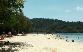 Crowded tropical beach at Sapi Island in Tunku Abdul Rahman Marine Park, showing white sand, turquoise ocean with swimmers, and a lush green mountain background during a Kota Kinabalu island hopping tour.