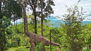 A life-sized sauropod dinosaur replica standing amidst lush green trees with Mount Kinabalu in the distant background at Jurassic Land Kiulu.