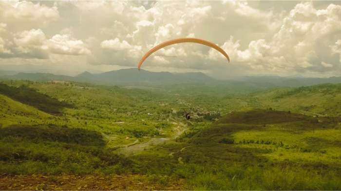 A paraglider soaring through a cloudy sky above the lush green hills and valleys of Ranau in Sabah.